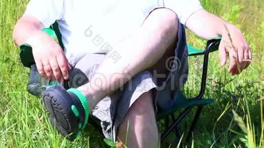 一个人坐在一片灯火通明的草地上的野餐椅上。 他休息，换腿。 草本植物和花朵从夏风中摇摆。