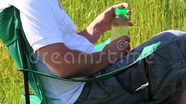 一个人坐在一片灯火通明的草地上的野餐椅上。 他休息一下，关上一瓶水，把它放在一个支架上，然后把他的手