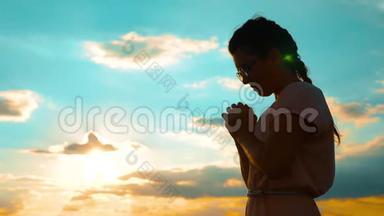 女孩在日落时双手交叉祈祷剪影。 慢动作视频。 女孩双手交叉祈祷祈求上帝