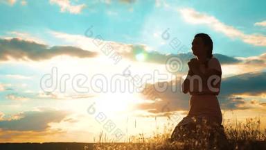 女孩在日落时双手交叉在生活方式祈祷剪影中。 女人跪着祈祷。 慢动作视频。 女孩