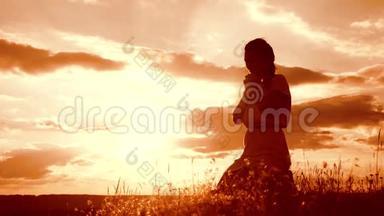 女孩在日落时双手交叉祈祷剪影。 女人跪着祈祷。 慢动作视频。 概念