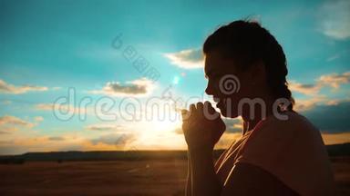 女孩在祈祷。 女孩在日落时交叉双手祈祷剪影。 慢动作视频。 女孩双手交叉祈祷