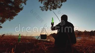 男子游客是啤酒坐在篝火晚会上，在大自然中露营，剪影，阳光日落。 户外男子剪影