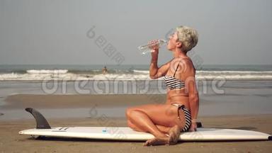 一个身材好的成熟女人坐在沙滩上，拿着冲浪板和喝水