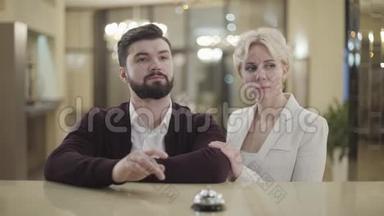 富有的白种人夫妇在酒店接待处按门铃和交谈。 金发女人和黑发男人的肖像