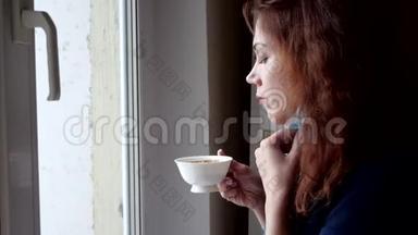 年轻女子戴着医疗面具喝咖啡，在隔离期间看着窗外。 社会距离
