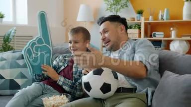 足球迷爸爸和孩子在家看电视比赛，手里拿着泡沫塑料球