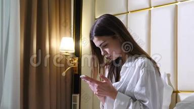 美丽的年轻女子坐在床上看着<strong>手机</strong>。 穿着男衬衫的女孩上网。 每<strong>日日</strong>报