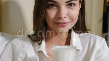 迷人的年轻女子微笑着看镜头喝牛奶或其他牛奶饮料的肖像。 一个漂亮的女孩