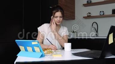 检疫期间厨房里的年轻女子。 女孩打电话，在笔记本上做笔记。 笔记本电脑和平板电脑