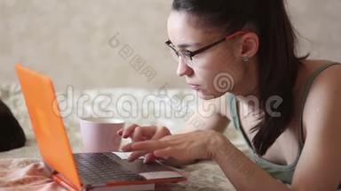 一个年轻迷人的女孩在家里躺在床上远程工作。 一个年轻可爱的女孩躺在床上用笔记本电脑工作。