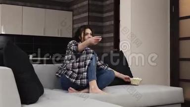 检疫期间厨房里的年轻女子。 快乐兴奋的女孩跳上沙发，用遥控器打开电视。 吃吃吃的