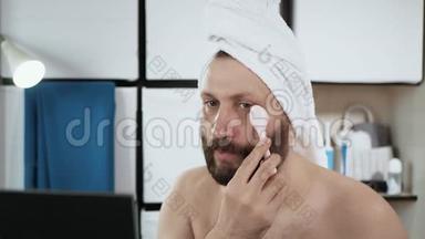 男人在他的眼睛下面做皮肤按摩。 变态，长胡子的男人头上戴毛巾在浴室用按摩器按摩皮肤
