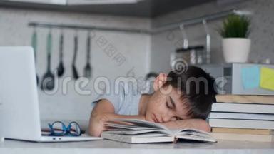 疲惫的小学生坐在笔记本电脑和课本旁边的书桌前做作业睡着了