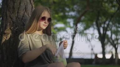 带着太阳镜的年轻女子坐在夏天公园的树旁，从棉网购物袋里拿苹果。 零废物