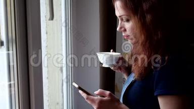 年轻女子手里拿<strong>着</strong>电话喝咖啡，在隔离期间<strong>看着窗外</strong>。 社会距离