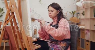 十几岁的孩子穿着艺术家的围裙，在她的房间里一只手拿着画笔。 患有唐氏综合症的女孩