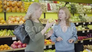 两个成熟的白种人家庭主妇在杂货店里闻着新鲜的黄梨，说话。 自信的女人讨论新鲜事物