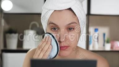 女孩用海绵擦她的脸。 浴室里漂亮的女人头上戴着毛巾，照镜子，洗脸
