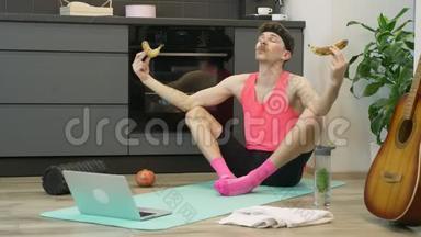 坐在莲花里的男人摆姿势，手里拿着香蕉在厨房的家中健身垫上。 带胡子和粉色衣服的有趣的年轻人