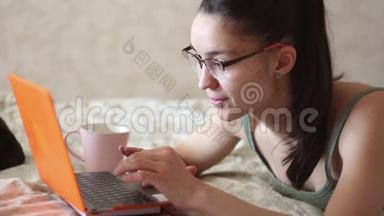 一个年轻迷人的女孩在家里躺在床上远程工作。 一个年轻可爱的女孩躺在床上用笔记本电脑工作。
