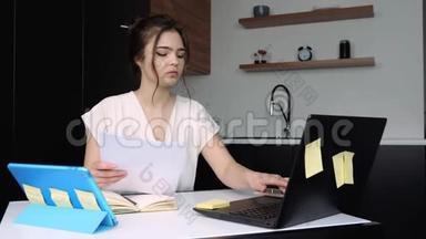 检疫期间厨房里的年轻女子。 严肃集中的女孩在笔记本电脑键盘上打字。 手拿文件