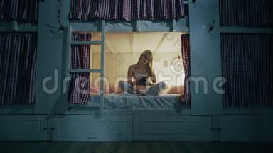 快乐微笑的女人坐在宿舍的床上，晚上用智能手机