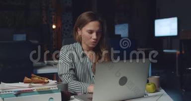 睡意朦胧的年轻女子深夜在办公室工作的肖像。 疲惫的女上班族用她的笔记本电脑打字