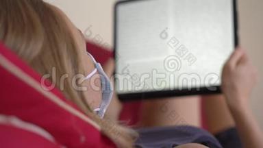 一位戴着医用口罩的年轻女子躺在沙发上看着一本电子书。 她在家吃冠状病毒