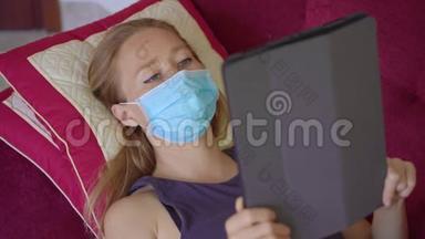 一位戴着医用<strong>口罩</strong>的<strong>年</strong>轻女子躺在沙发上看着一本电子书。 她在家吃冠状病毒