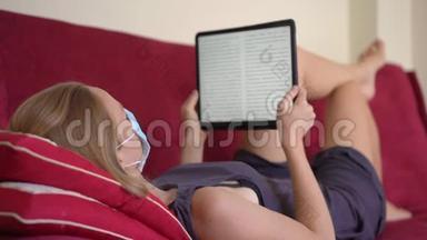 一位戴着医用<strong>口罩</strong>的<strong>年</strong>轻女子躺在沙发上看着一本电子书。 她在家吃冠状病毒