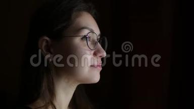 戴眼镜的年轻女子从一间昏暗的房间里向窗外沉思地看着