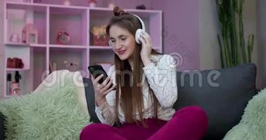女孩正在通过耳机听音乐，并使用智能手机在家里的沙发上放松，玩得很开心。 小玩意，开心