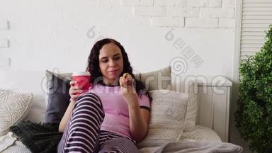一个年轻的女人在床上吃热狗和喝咖啡。 在床上吃早餐。 把食物送到你家。 女人吃东西
