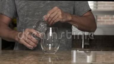穿着灰色T恤的年轻人在一个漂亮时尚的厨房里用热水瓶或塑料瓶把水倒入玻璃杯中