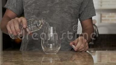 穿着灰色T恤的年轻人在一个漂亮时尚的厨房里用<strong>热水</strong>瓶或塑料瓶把水<strong>倒</strong>入玻璃杯中