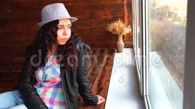 戴着帽子和皮夹克的年轻美女坐在窗户旁。 从窗户向外看的漂亮女人的肖像