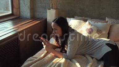 穿着休闲服躺在床上的漂亮年轻女人。 成人微笑的女士在手机里<strong>翻阅</strong>各种新闻。