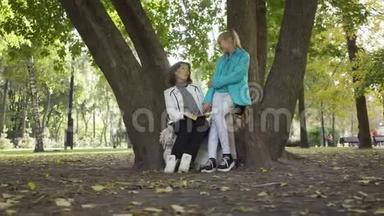 两个白种人妇女坐在秋天公园的树上讨论这本书。 漂亮的黑发女人解释