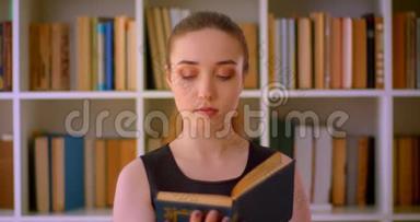 年轻成功的红头发女学生在室内图书馆阅读一本带有书架的书