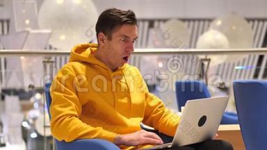 年轻人坐在咖啡厅时，在笔记本电脑上想着震惊。 穿着黄色连帽衫的男人被他在电脑上看到的东西震惊了