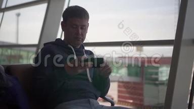 美国男子坐在机场，坐在帕诺曼的窗户，椅子上，看手机上的应用程序