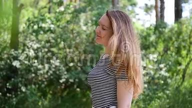 年轻的棕色头发的女人，穿着条纹t恤，站在阳光下，迎着绿色的树叶，风吹起她的头发。 漂亮姑娘