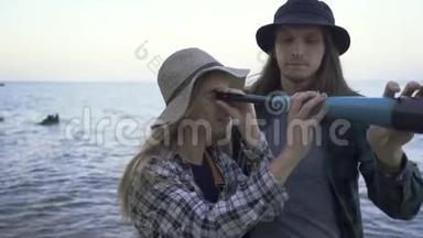 年轻的高加索游客夫妇穿着衬衫和帽子，看着望远镜，在<strong>大海</strong>或湖泊的<strong>背景</strong>下聊天。