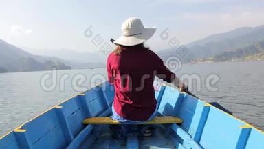 穿着红色衬衫，戴着帽子的女孩坐在一艘蓝色的木船上，手里拿着桨，从后面看着湖，靠在后面