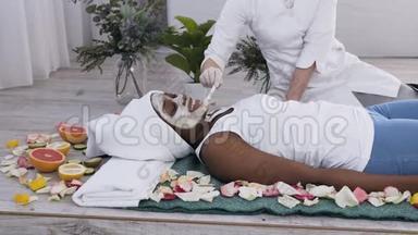 年轻漂亮的非洲裔美国妇女，头上有白色毛巾，躺在沙发上放松，美容师为她做面部护理