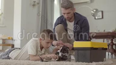 <strong>父亲和儿子在家里</strong>的地板上铺着毛茸茸的地毯，固定着破碎的虚拟现实头盔特写。 孩子