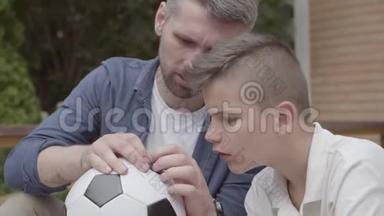 父亲和儿子坐在门廊上的肖像，手里拿着一个泄气的足球特写。 他爸爸在帮助