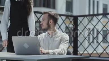 大胡子的年轻人坐在笔记本电脑前的露台上的桌子旁工作。 可爱的女孩来看着