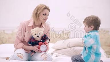 年轻的妈妈带着可爱的小儿子和泰迪熊<strong>坐在</strong>家里的<strong>床上</strong>。 友好的家庭，一位年轻的母亲<strong>坐在</strong>旁边